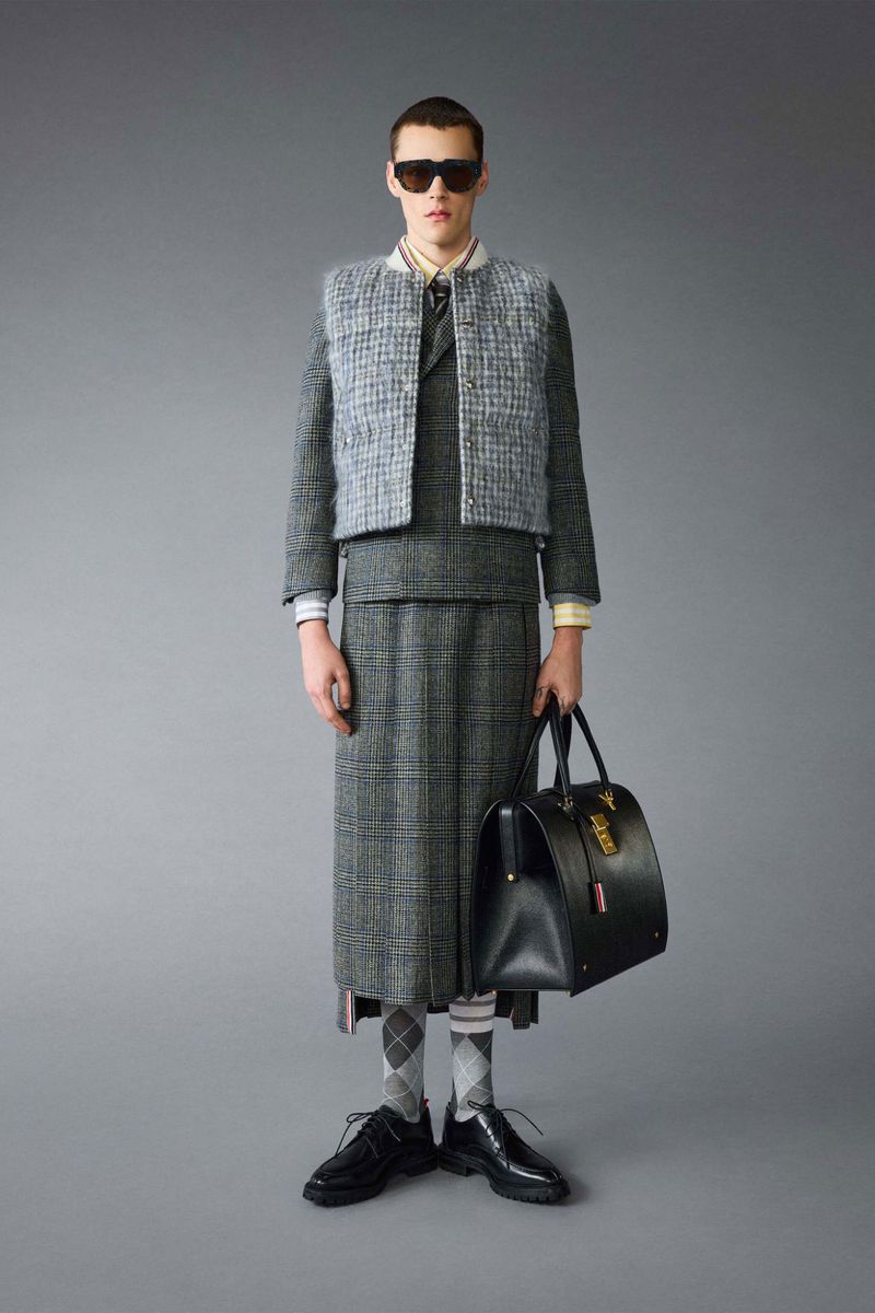Man poses in grey tweed skirt suit with vest, white collared shirt, dark sunglasses, black dress shoes, socks and black leather doctor's bag.