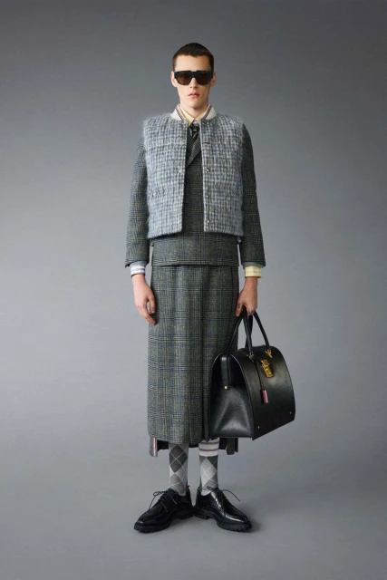 Man poses in grey tweed skirt suit with vest, white collared shirt, dark sunglasses, black dress shoes, socks and black leather doctor's bag.