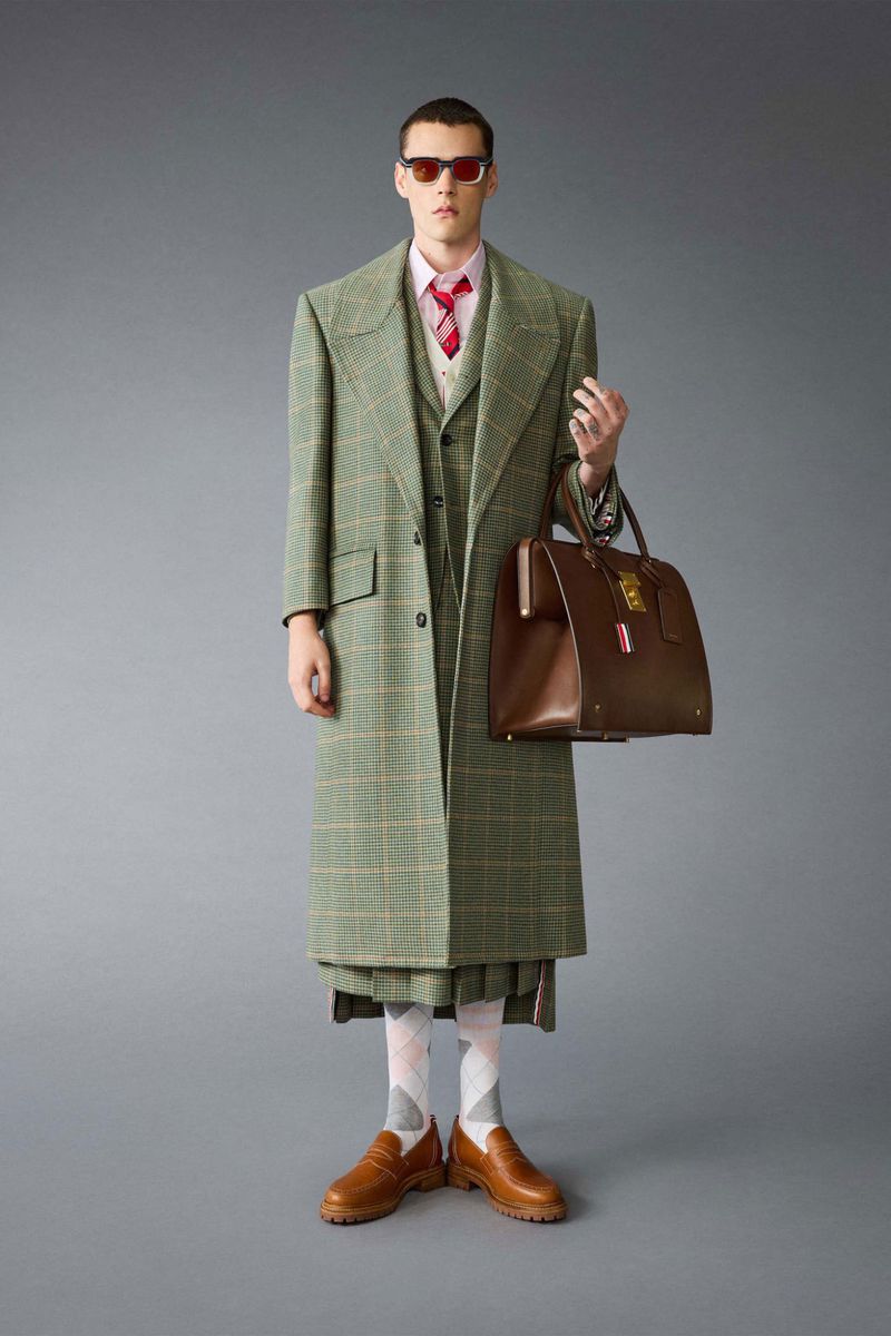 A man in a green plaid suit, button-down shirt, and tie holds a brown leather bag against a grey studio backdrop, showcasing men's fashion.