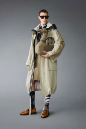 Model in layered tan coat holding a goose-shaped bag against a neutral grey studio backdrop
