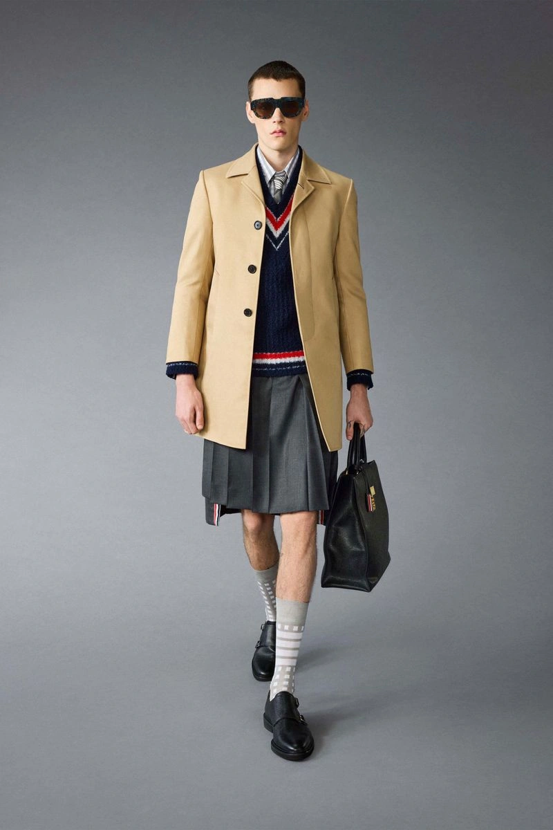 Model wearing a tan coat, pleated grey skirt, and holding a black handbag in a studio