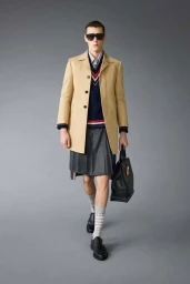 Model wearing a tan coat, pleated grey skirt, and holding a black handbag in a studio