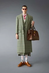 Model wearing a plaid green coat and suit holding a brown leather bag against a grey seamless background