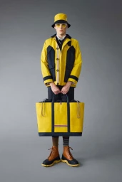 Male model in yellow jacket and bucket hat holding a large yellow tote bag against grey seamless background
