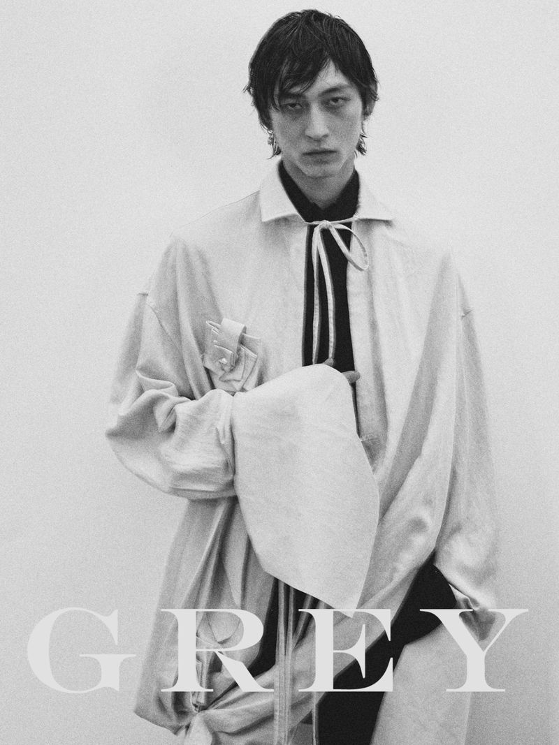 Black and white fashion portrait of an adult male wearing a light colored coat and black shirt with the word 'GREY' visible.