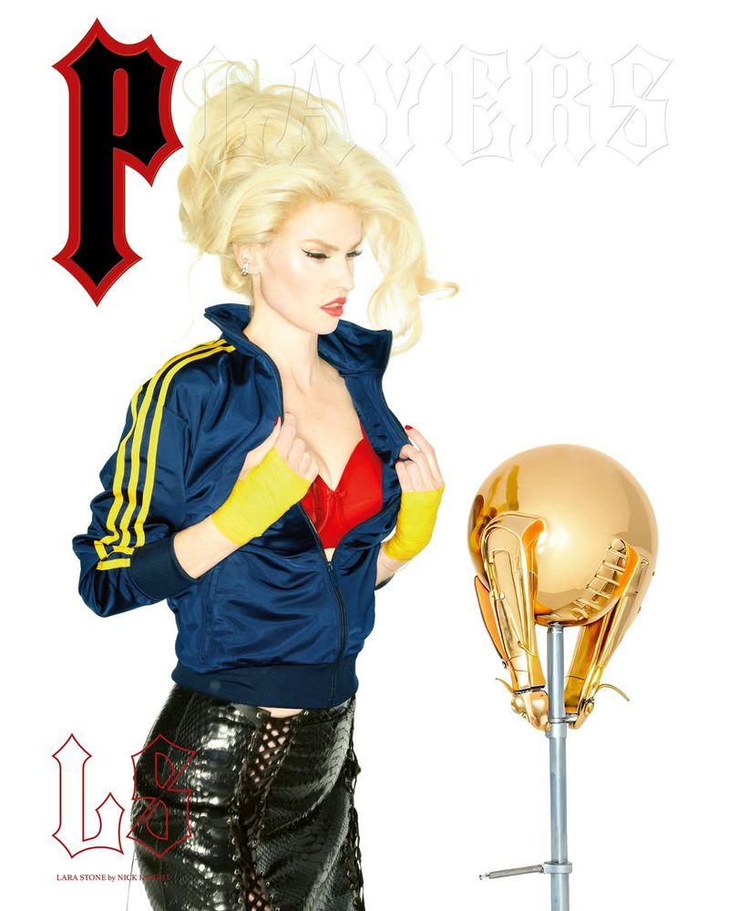 Blonde woman in a jacket poses next to a golden football helmet on a white background in a fashion shot. Red bra is visible.