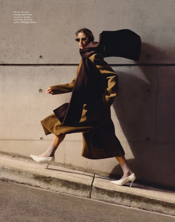 Fashion editorial shot: A stylish woman in a brown coat, scarf, and white heels walking against a concrete wall. Editorial fashion.
