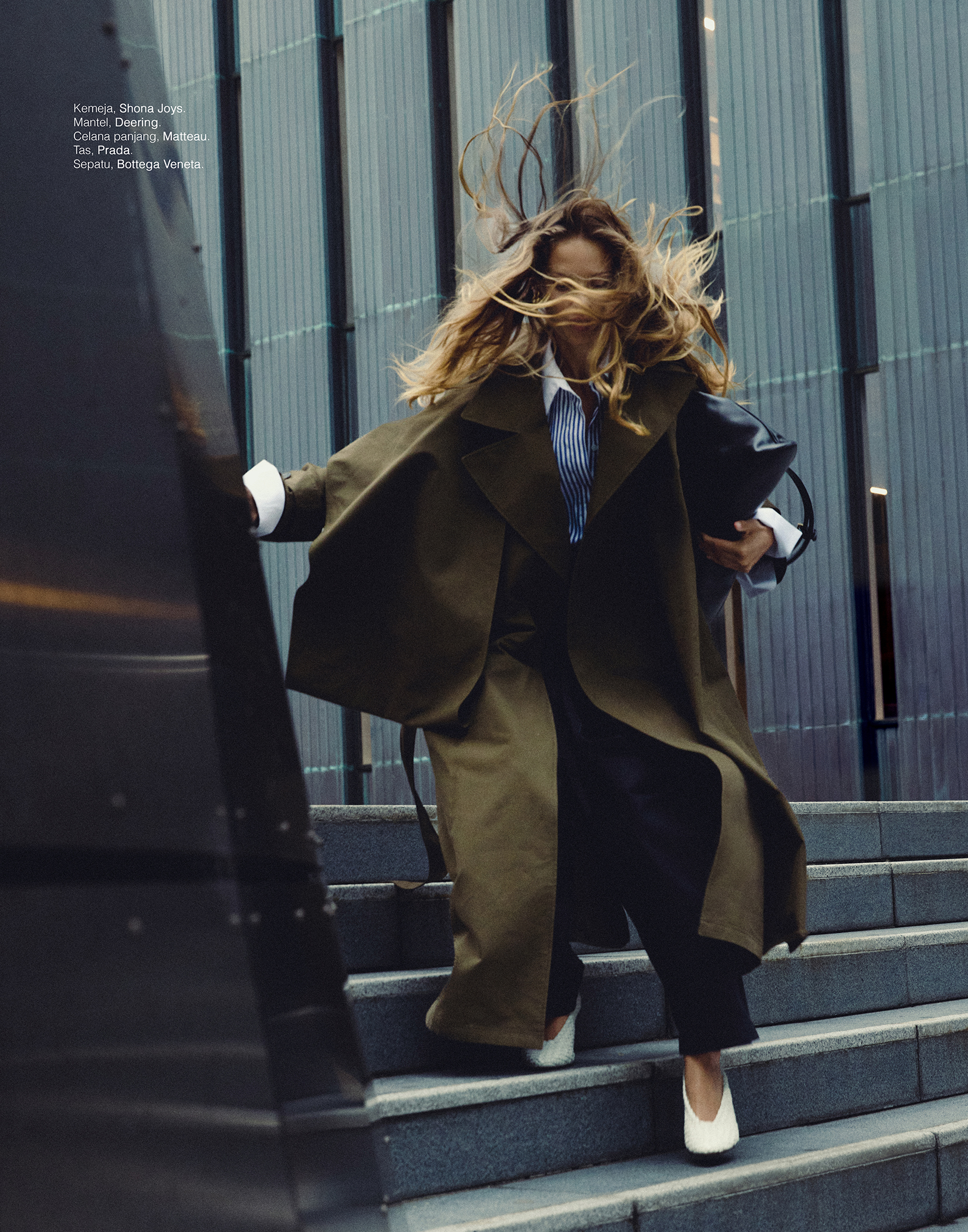 Fashionable woman in oversized olive coat and pants strides on stairs, her hair blowing in the wind, capturing a dynamic editorial moment.
