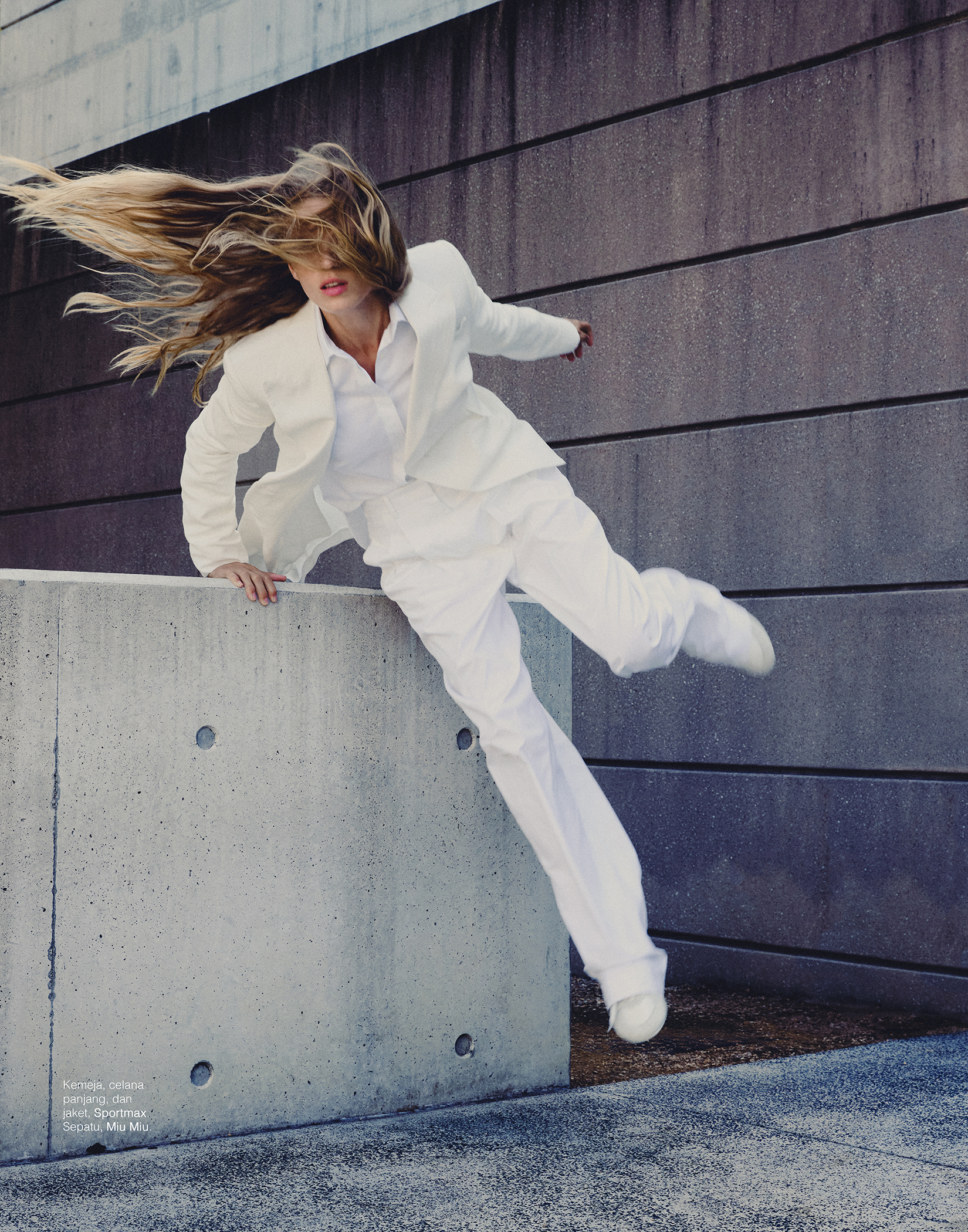 Fashionable woman in a white suit leaping off a concrete block in an urban setting, her blonde hair flying, and an active pose.