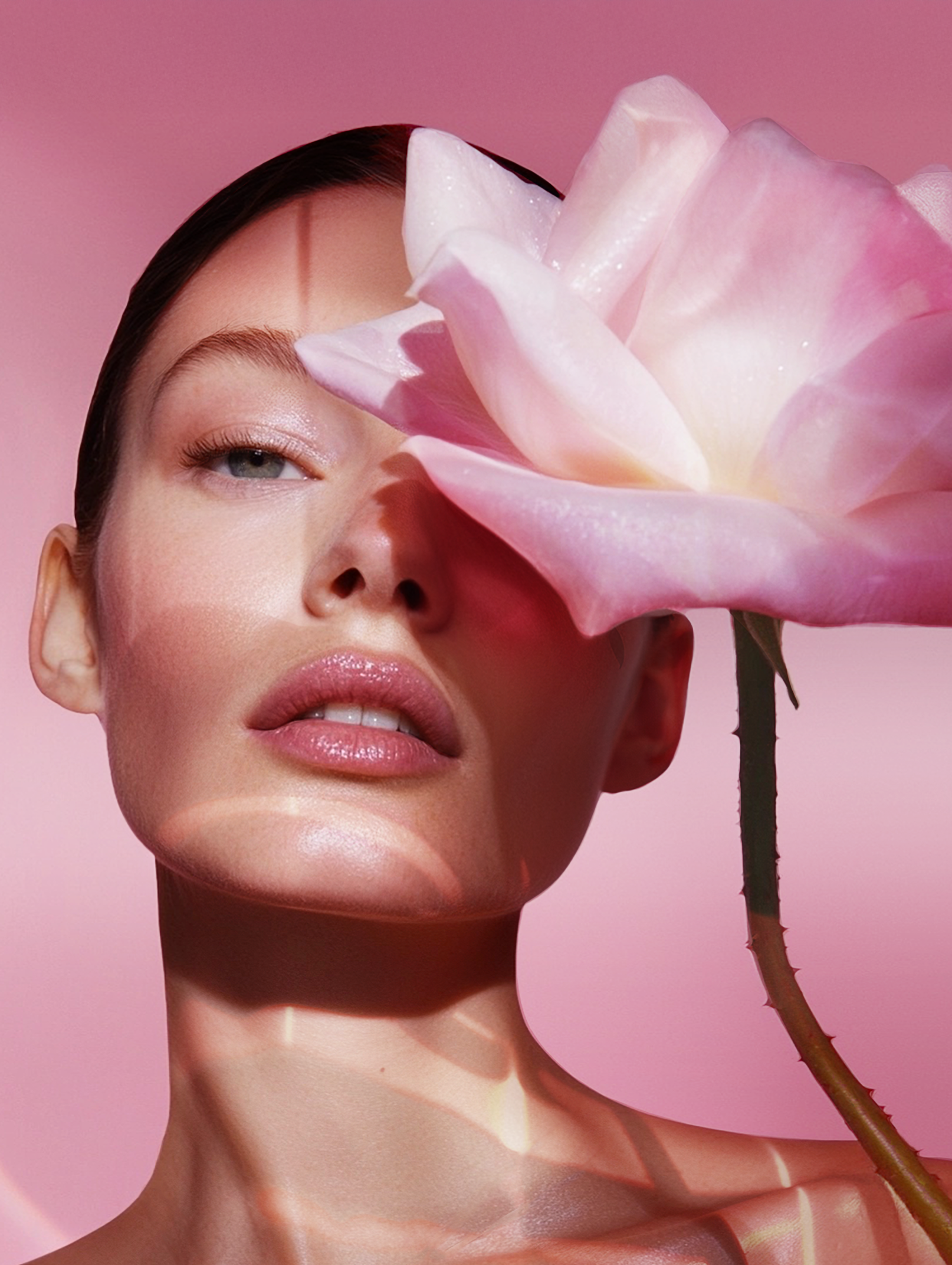 Close-up of a woman with fair skin partially obscured by a large, pale pink rose, against a soft pink backdrop with reflected light.