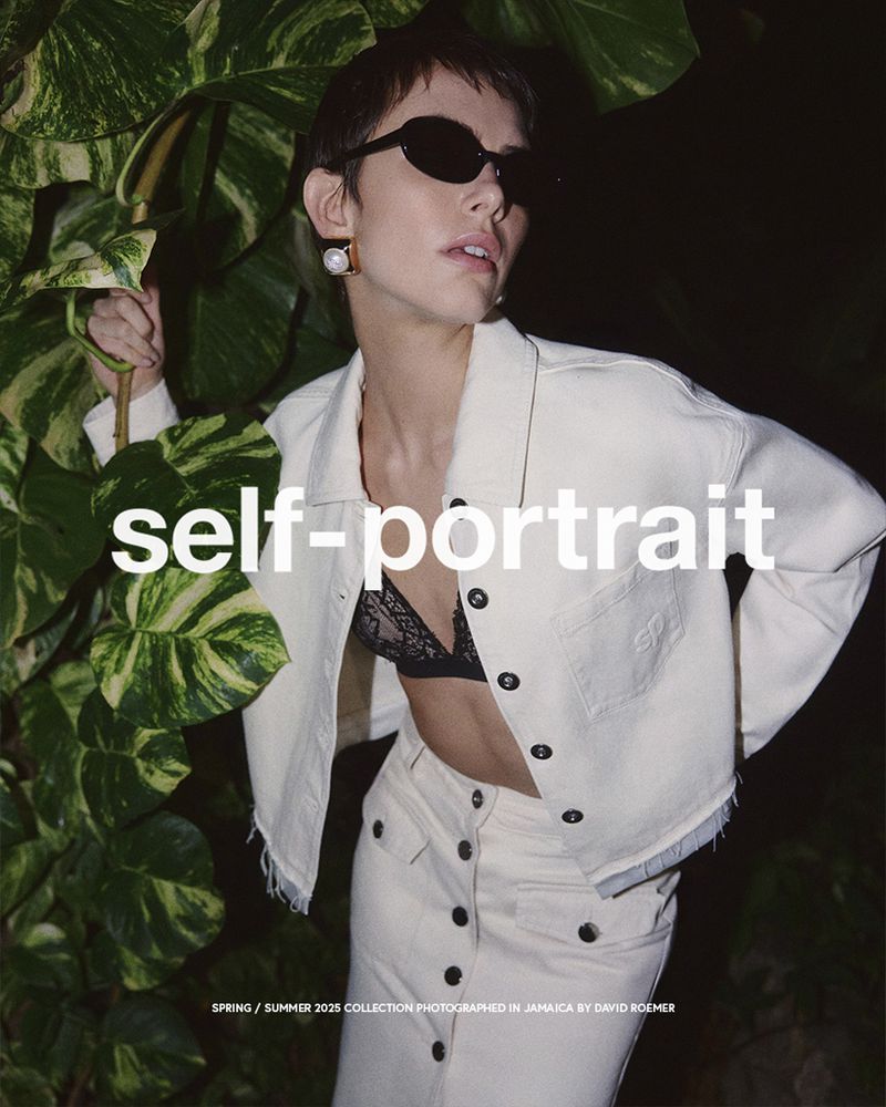 Fashion shot of a woman wearing a cream-colored denim jacket, skirt, and black lace bra, posing with tropical foliage.