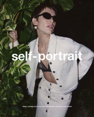Fashion shot of a woman wearing a cream-colored denim jacket, skirt, and black lace bra, posing with tropical foliage.