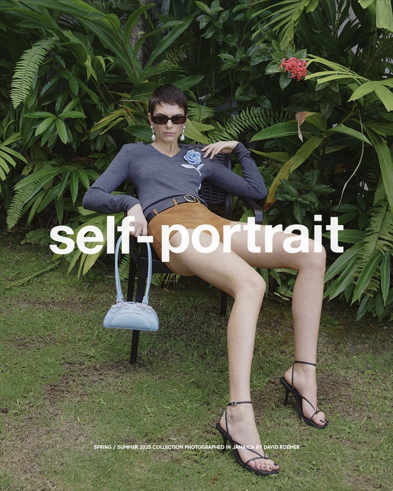 A slender woman poses on a chair outdoors wearing a grey sweater, a tan skirt, and strappy heeled sandals, with greenery as backdrop.