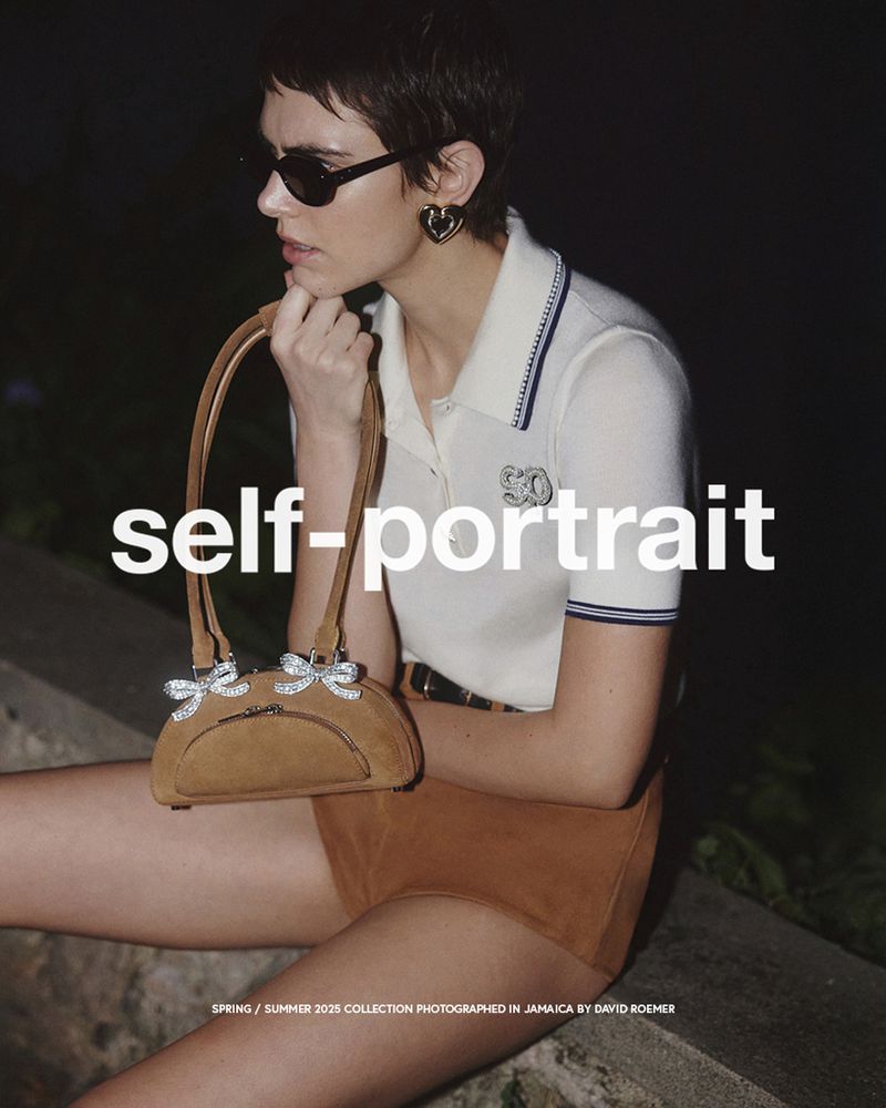 Fashion shot of a woman with a suede purse wearing a white collared shirt and tan shorts with black sunglasses, shot in an outdoor setting.
