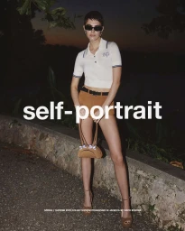 Model wearing white polo shirt, brown shorts, and sunglasses holding a handbag outdoors at dusk