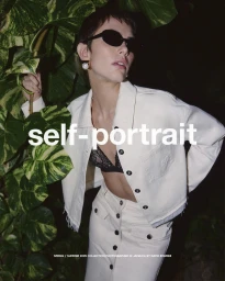 Model wearing dark sunglasses and an off-white denim jacket posing amidst lush tropical foliage