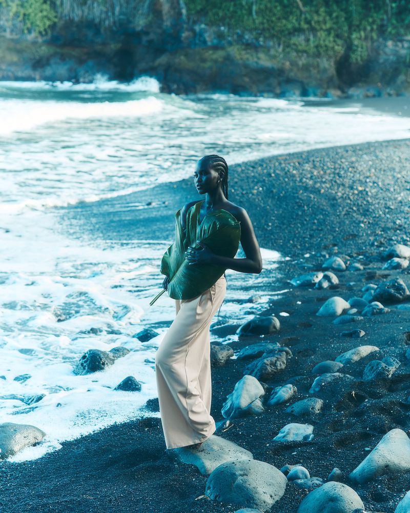 A dark-skinned woman with green textile, standing on a black sand beach near waves, wears tan pants in a portrait-style fashion shot.