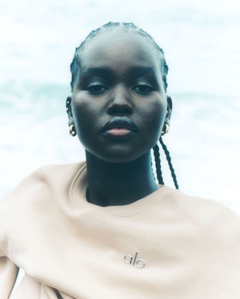 Portrait of a young woman with dark skin, braided hair, gold earrings, and a beige sweatshirt posing against a blurred, light blue background.