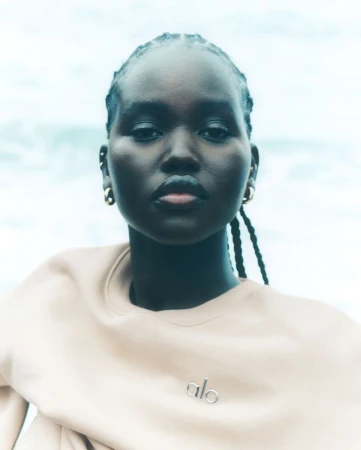 Portrait of a young woman with dark skin, braided hair, gold earrings, and a beige sweatshirt posing against a blurred, light blue background.