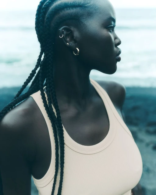 Portrait of an adult woman with dark skin wearing a light-colored tank top with braided hair against a blurred ocean background.