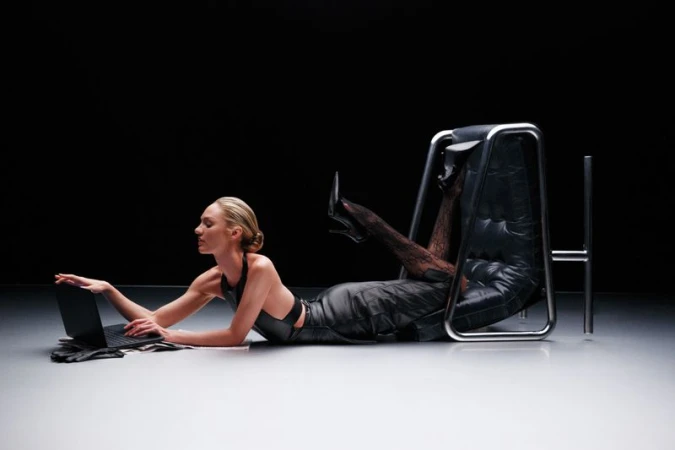 Woman in black outfit reclines with laptop next to a modern chair in a stark studio with black backdrop and light grey floor.