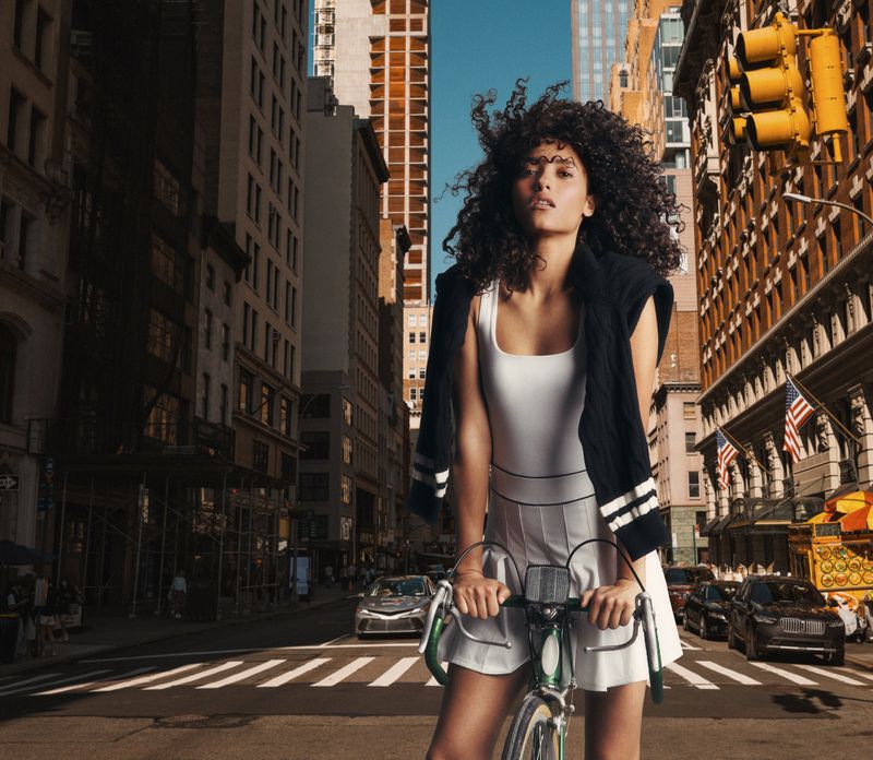 A curly-haired woman in a white dress rides a bicycle on a sunny urban street lined with tall buildings and yellow traffic lights.
