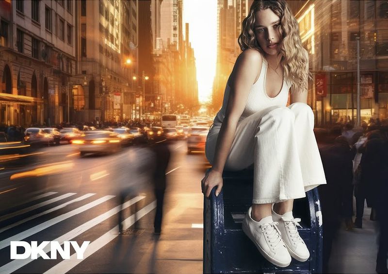 Fashion shot of a woman in white pants and top sitting on a blue box against an urban background with blurred cars and people.