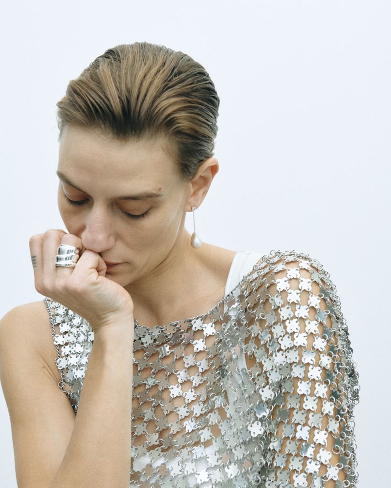 A fair-skinned adult woman poses indoors, wearing a metallic top and silver ring, against a white backdrop in a medium shot.