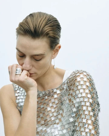 A fair-skinned adult woman poses indoors, wearing a metallic top and silver ring, against a white backdrop in a medium shot.