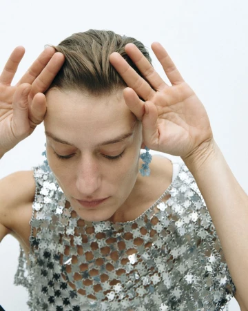 Woman poses with hands near her head in a studio shot, wearing a silver sequin top and blue earrings against a white backdrop.