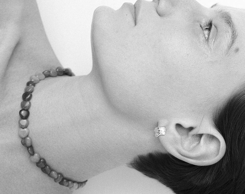 Monochrome portrait of a woman with a beaded necklace and ear cuff, lying down against a white background, focusing on jewelry and skin tone.