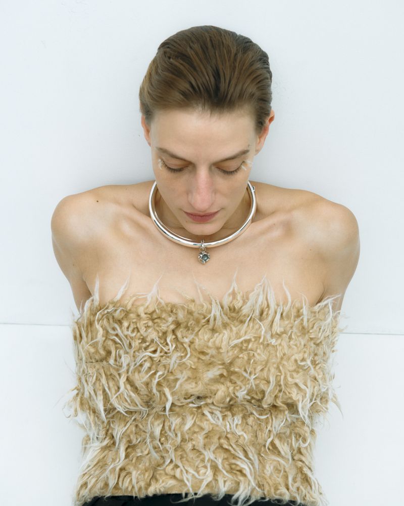 Woman in furry tan top and silver choker poses against a white background, looking down in a fashion studio shot with soft lighting.
