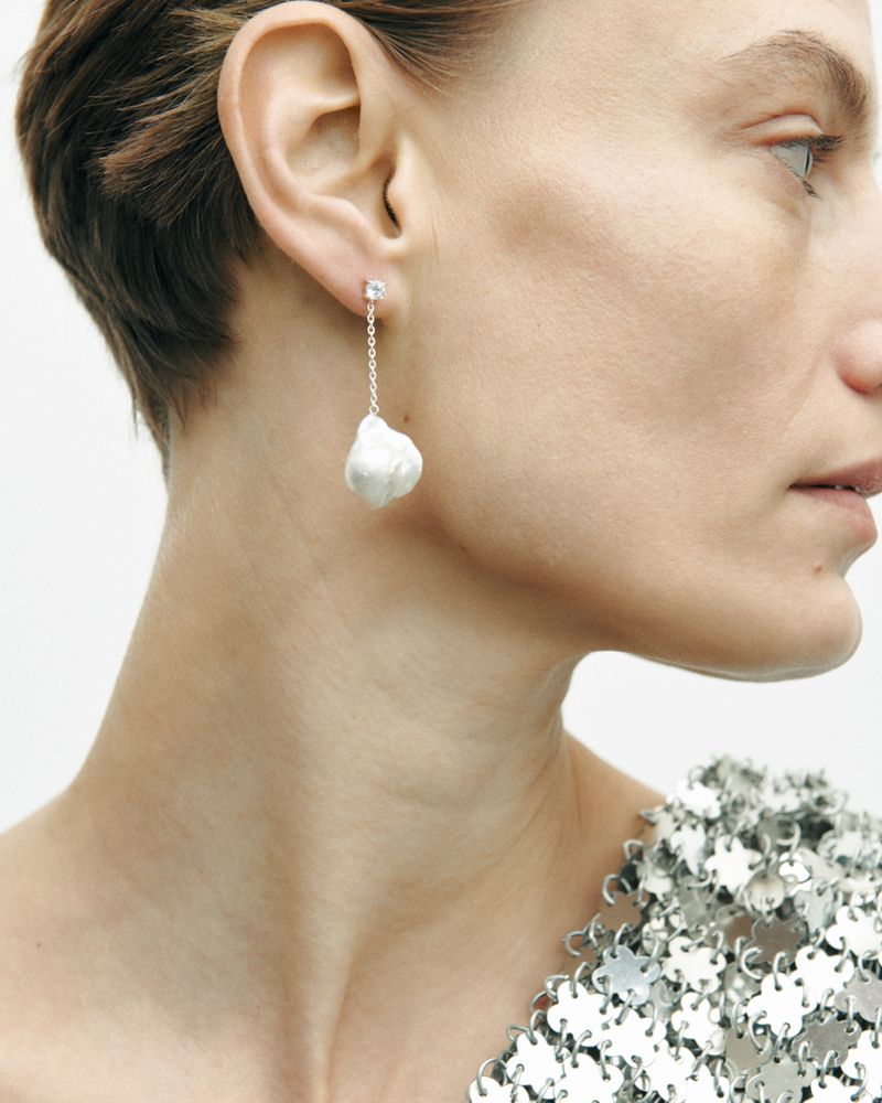 Close-up studio shot of an adult female model wearing a silver metallic dress and baroque pearl earring, in a portrait orientation.