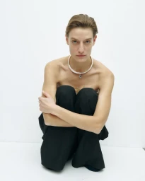 Model wearing a silver choker necklace and black trousers crouching against a seamless white background