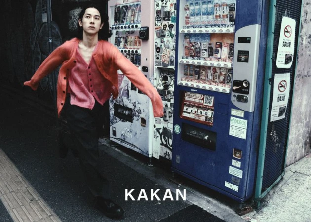A slim Asian man in a pink cardigan, dancing in an urban alley next to vending machines, captured in a vibrant street-style photograph.