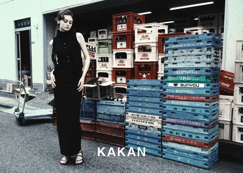 Adult woman in a black dress poses in an urban setting with stacks of blue and white crates. Outdoor fashion portrait.