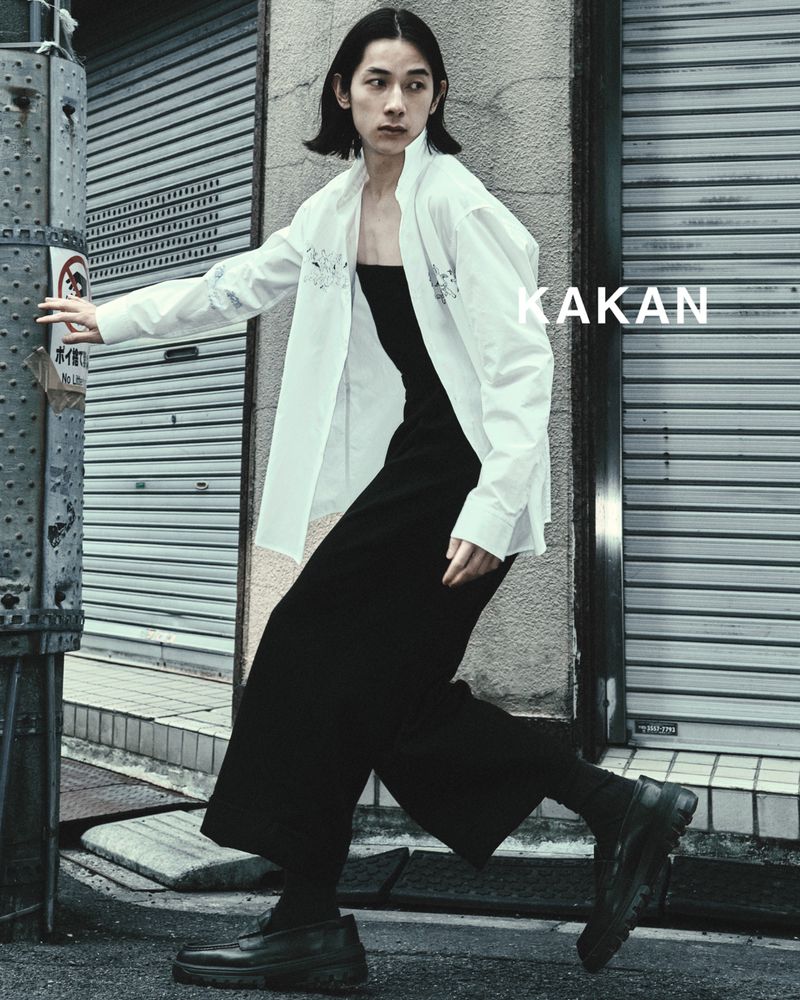 Asian male model in a white shirt and black culottes poses in an urban setting with a weathered metal wall and street pole.