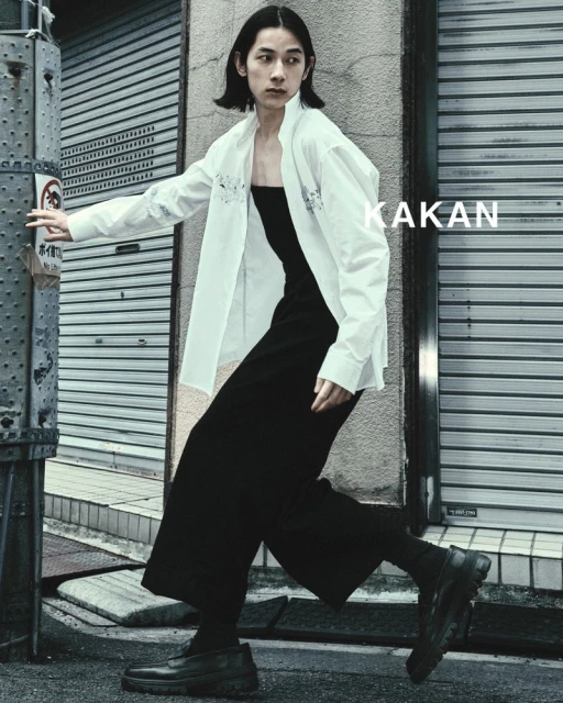 Asian male model in a white shirt and black culottes poses in an urban setting with a weathered metal wall and street pole.