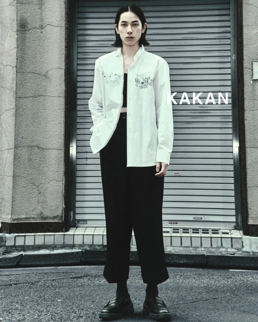 A slender person stands in an urban setting, wearing an unbuttoned white shirt with embroidery over a black top and black pants.