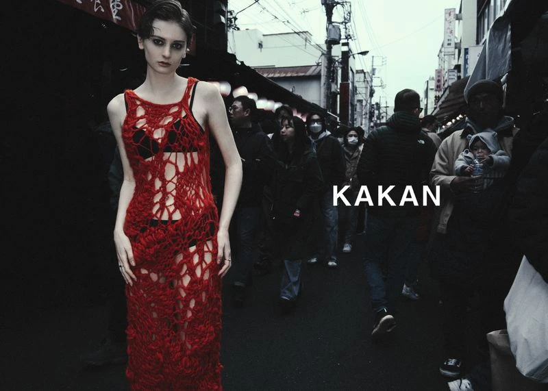 Fashion shot of a woman in a red crochet dress standing in a crowded urban street with overcast lighting and blurred figures behind her.