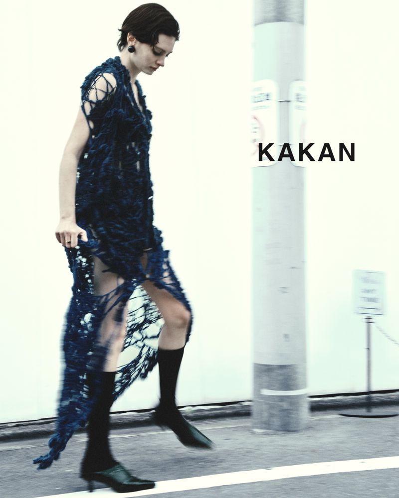 A slender female model in a unique, dark blue woven dress strides on a city street with KAKAN logo beside her, styled with socks and heels.