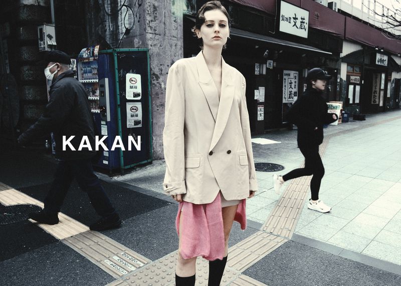 A woman in a beige jacket stands on a city street with a runner and a passerby in the background, creating an urban fashion scene.