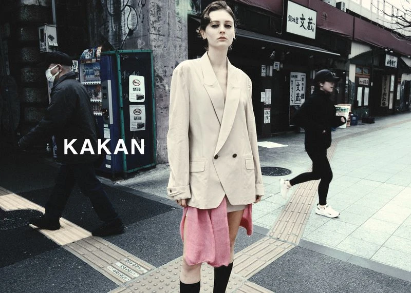 A woman in a beige jacket stands on a city street with a runner and a passerby in the background, creating an urban fashion scene.