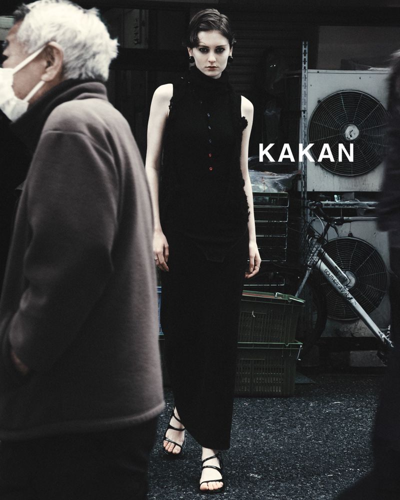 Woman in a sleeveless black dress walks on a city street as people pass by; industrial equipment and a black bicycle are in the background.