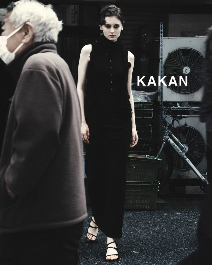 Model in black sleeveless top and long skirt walking on an urban street with blurred figures in foreground and background.
