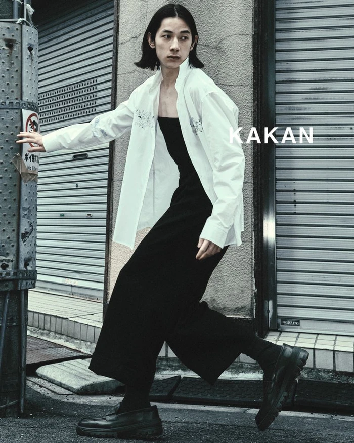Model wearing a white shirt and black trousers stepping past metal shutter doors outdoors