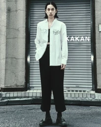 Model standing in front of a closed roller shutter door wearing a white shirt and black trousers for KAKAN SS25