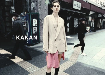 Model wearing an oversized beige blazer and pink fabric detail on a city street with pedestrians walking in the background.