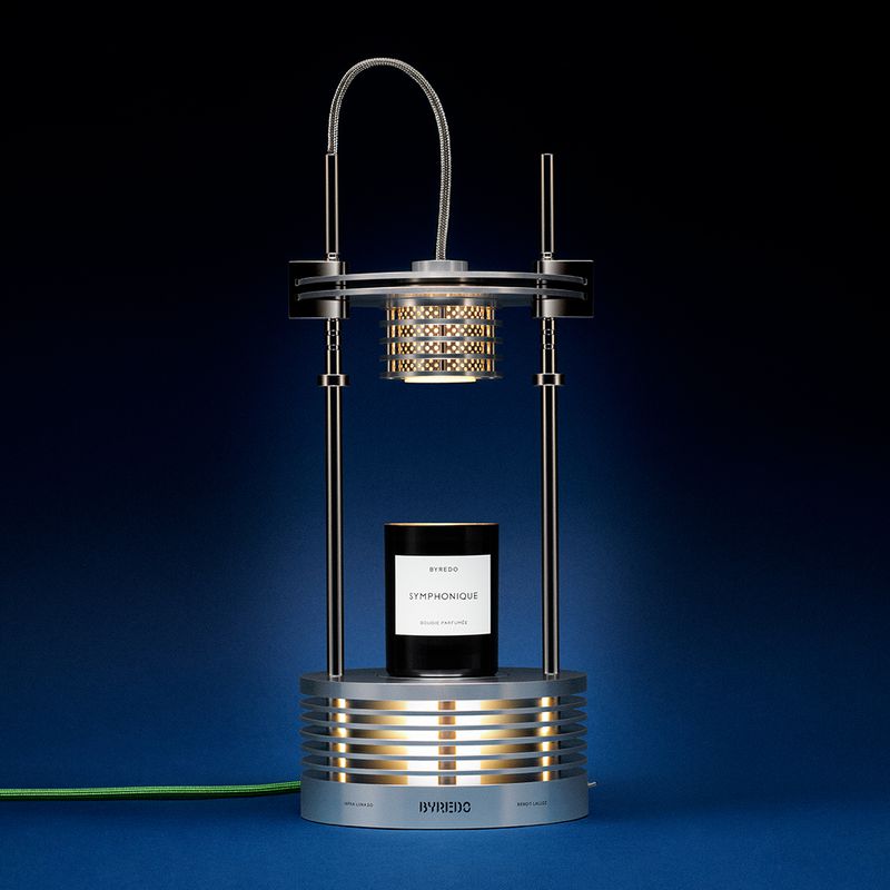 Silver Byredo candle holder with a black Byredo candle, studio shot against a blue backdrop, well-lit product photography.