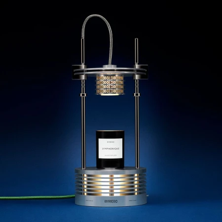 Silver Byredo candle holder with a black Byredo candle, studio shot against a blue backdrop, well-lit product photography.
